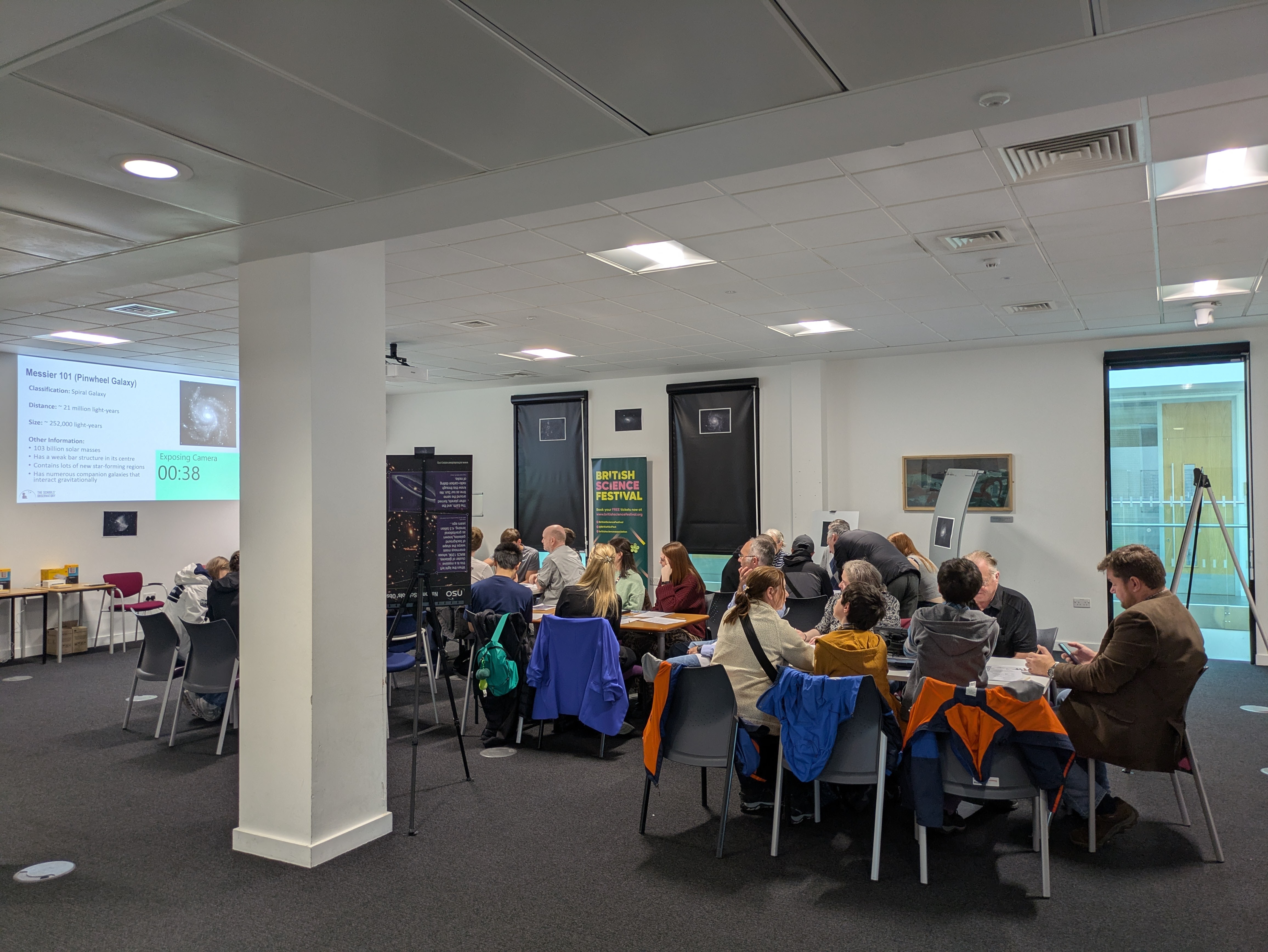 a photograph of a group of about 20 people taking part in a session. The people are split into groups across different tables and there is a presentation slide with information about the Pin Wheel galaxy